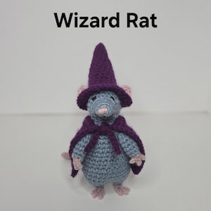 May include: A crocheted gray rat figurine wearing a purple pointed wizard hat and a matching purple cape tied at the neck. The rat has small pink ears, a pink nose, and pink feet. The text Wizard Rat is printed in black above the figurine.