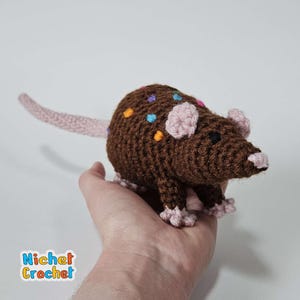 May include: A handmade crochet rat in shades of brown, with colorful sprinkles on its back. The rat has pink ears, a pink nose, and a long, pink tail. The crochet animal is held in a hand.
