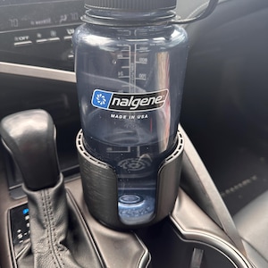 May include: A clear, dark blue Nalgene water bottle with a black lid and handle sits in a black cup holder. The bottle has measurement markings and the Nalgene logo. The text "MADE IN USA" is visible on the bottle.
