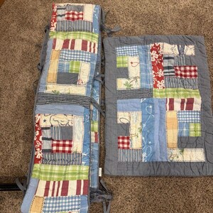 May include: A patchwork crib bumper and quilt set. The bumper is a long, rectangular shape with ties, and the quilt is square. Both pieces feature a variety of fabric patterns in blue, red, green, and white, including stripes, checks, and floral prints.