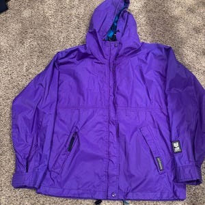 May include: A vibrant purple hooded jacket with a full zipper closure and two front pockets. The jacket features a drawstring at the bottom hem and a small logo patch on the sleeve. The interior lining of the hood has a blue and white pattern.