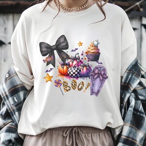 May include: White t-shirt featuring a watercolor Halloween design. The design includes pumpkins, a black bow, a cupcake with a ghost, and the word "BOO". Other elements include bats, stars, and a coffin.