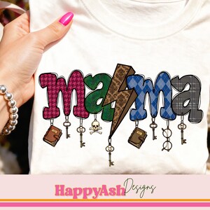 Op de afbeelding: Wit T-shirt met het woord "mama" in een kleurrijk, patroonontwerp. De letters zijn versierd met pied-de-poule, groen, blauw geruit en een bliksemschicht. Kleine bedels van sleutels, boeken en brillen hangen eronder.