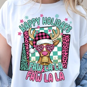 Puede incluir: Camiseta blanca con un gráfico festivo. El diseño presenta un reno con gafas de sol con estampado de leopardo en forma de corazón y un gorro a cuadros. El texto dice "Happy Holidays" y "Faw La La" en una fuente lúdica. Acentos rosas y turquesas.