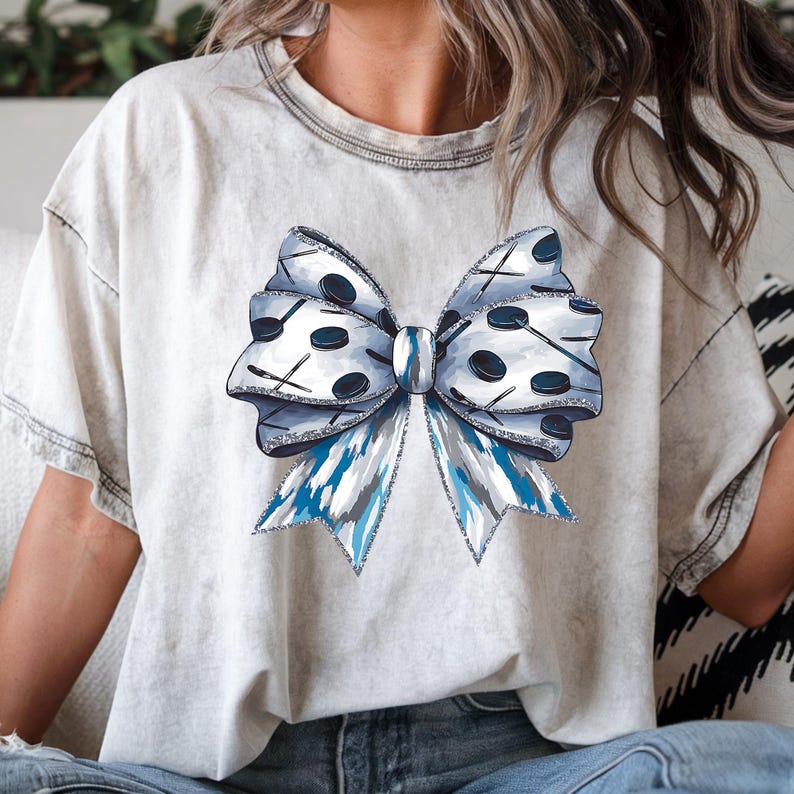 Puede incluir: Una camiseta blanca con un dise&ntilde;o de lazo azul y blanco. El lazo est&aacute; hecho de discos y palos de hockey.