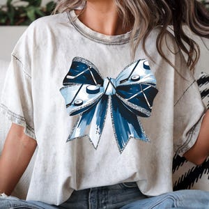 Puede incluir: Una camiseta blanca con un dise&ntilde;o de lazo azul y plateado con purpurina que presenta palos de hockey y discos.