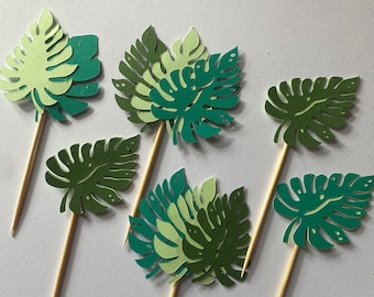 May include: Six green and white paper leaf cupcake toppers with wooden sticks. The leaves are in various shapes and sizes, with some having a layered look.