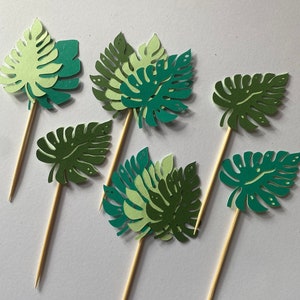 May include: Six green and white paper leaf cupcake toppers with wooden sticks. The leaves are in various shapes and sizes, with some having a layered look.