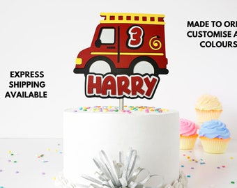 May include: Red fire truck cake topper with the name "HARRY" and the number "3" on the side. The topper is on a white cake with colorful sprinkles.