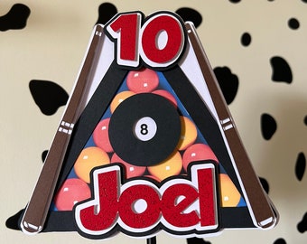 May include: A red and white cake topper with a pool table design. The table has a black felt surface with a black 8 ball and red and yellow billiard balls. The topper also includes the number 10 and the name Joel in red.