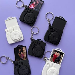 May include: A selection of camera-shaped keyrings in black and white, some with photos displayed. The keyrings feature a heart design and a silver keyring. The background is a solid purple colour.