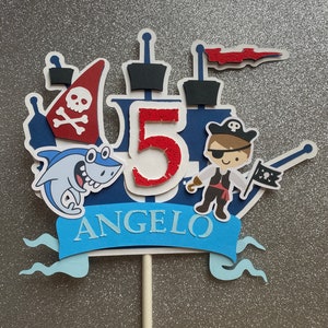 Op de afbeelding: Een blauwe en rode piratenboot-taarttopper met een haai, een piraat en het getal 5. De topper bevat ook een blauwe banner met de naam "ANGELO" erop.