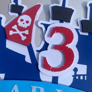 Op de afbeelding: Een rode en witte piratenboot-taarttopper met een schedel en gekruiste beenderen op het zeil en het nummer 3 aan de zijkant. De taarttopper staat op een blauwe achtergrond met het woord "ARIO" in wit.