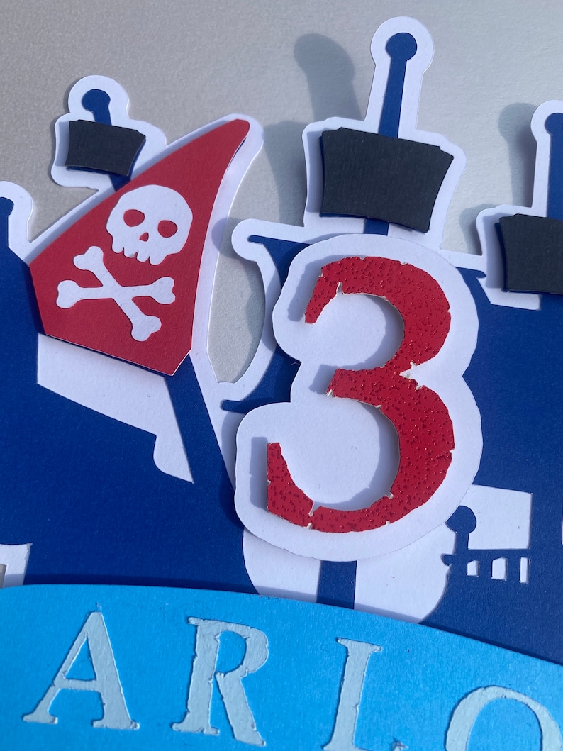 Op de afbeelding: Een rode en witte piratenboot-taarttopper met een schedel en gekruiste beenderen op het zeil en het nummer 3 aan de zijkant. De taarttopper staat op een blauwe achtergrond met het woord "ARIO" in wit.
