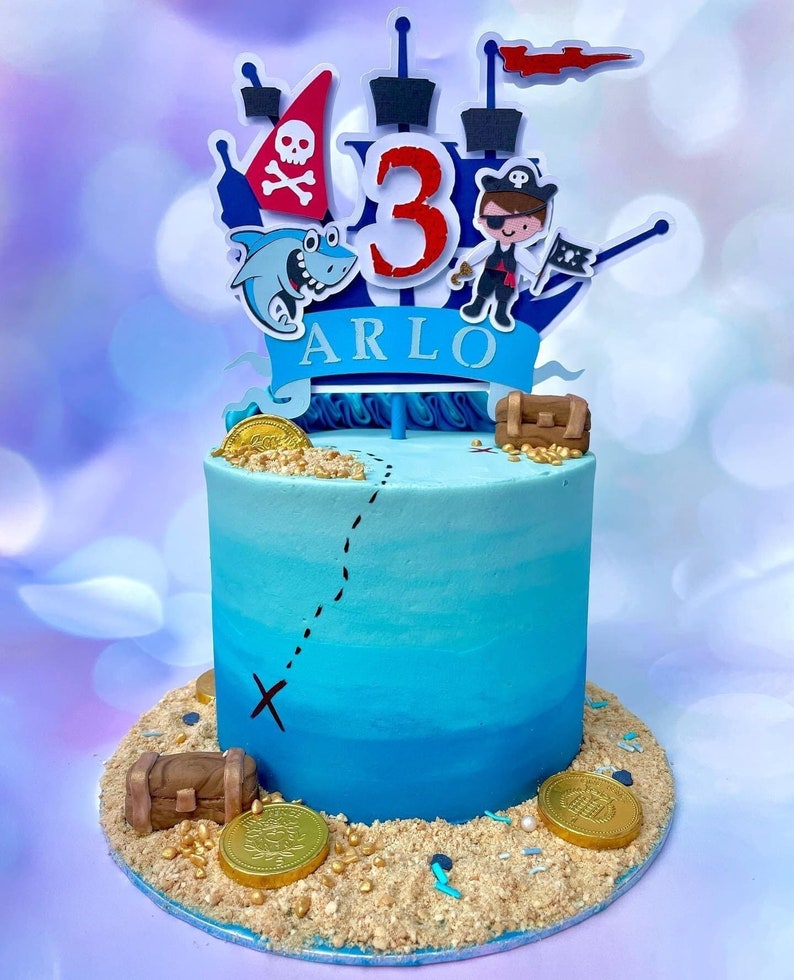 Op de afbeelding: Een blauwe en witte taart versierd met een piratenschip, een haai en gouden munten. De taart is versierd met een bord waarop "Arlo 3" staat.