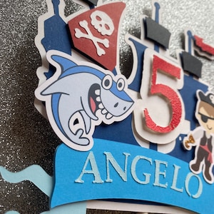 Op de afbeelding: Een blauwe en rode piratenboot-taarttopper met een haai, een piraat en het nummer 5. De topper bevat ook de naam "Angelo" in blauwe letters.