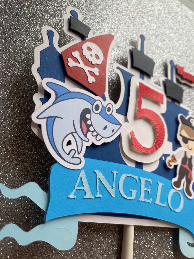Op de afbeelding: Een blauwe en rode piratenboot-taarttopper met een haai, een piraat en het nummer 5. De topper bevat ook de naam "Angelo" in blauwe letters.