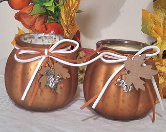 Puede incluir: Dos candelabros redondos de color cobre con cintas blancas y naranjas atadas en la parte superior. Cada candelabro tiene un dije de ardilla plateada y una etiqueta en forma de hoja marrón. El fondo incluye follaje otoñal.