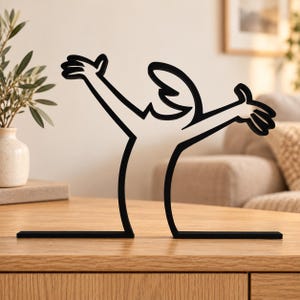 Puede incluir: Una escultura de metal negro que presenta un dibujo de línea continua y estilizada de una figura humana con los brazos levantados en un gesto de celebración. La figura se muestra en una forma de silueta simple y audaz sobre una base plana.