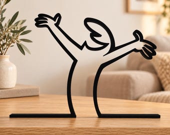 May include: A black metal sculpture featuring a stylized, continuous line drawing of a human figure with arms raised high in a gesture of celebration. The figure is depicted in a simple, bold silhouette form, standing on a flat base.