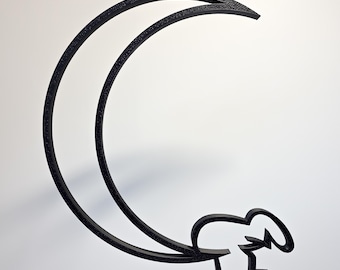 May include: Black metal wall art featuring a crescent moon design with a stylized figure hanging from it. The artwork is suspended from a thin wire, creating a floating effect. The piece has a textured surface and is set against a white background.