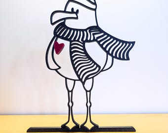 May include: A black and white seagull figurine wearing a striped scarf and hat, with a red heart on its chest. The seagull is standing on a black base, and the background is a light color.