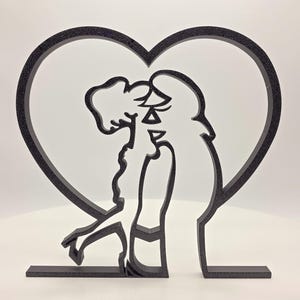 May include: A black sculpture featuring two figures embracing within a heart-shaped outline. The artwork is a solid, three-dimensional piece, possibly made of a matte material, and stands on a rectangular base.