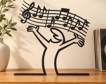 May include: A black metal sculpture featuring a stylized human figure with arms raised, supporting a curved musical staff with notes and a treble clef. The piece is constructed from a single continuous line design, standing on a flat base.
