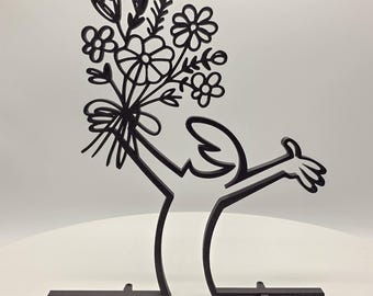 May include: Black metal sculpture of a person holding a bouquet of flowers. The sculpture is on a black metal base with three legs.