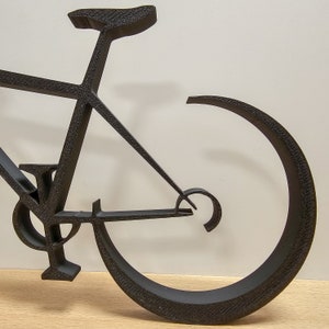 Puede incluir: Escultura de bicicleta de metal negro con un marco curvo y una sola rueda. La escultura est&aacute; hecha de metal delgado y tiene un dise&ntilde;o minimalista.