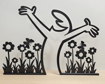May include: Black silhouette of a person with outstretched arms standing in a field of flowers. The person is made of wood and the flowers are cutouts. The flowers are in a field of grass.