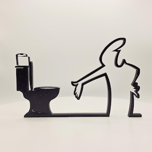 Könnte beinhalten: Ein schwarzes, flaches, silhouettenartiges Dekorationsobjekt, das eine Toilette mit einer Toilettenpapierrolle darauf zeigt, neben einer stilisierten menschlichen Figur mit ausgestrecktem Arm, die auf die Toilette zeigt, alles verbunden durch eine Basis.