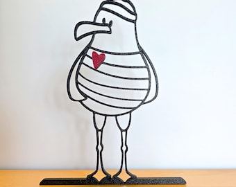 May include: A black and white seagull sculpture with a red heart on its chest. The seagull wears a striped shirt and a striped hat. The sculpture is standing on a black base and is made of a glittery material.