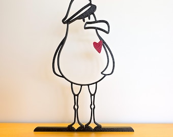 May include: A black metal seagull sculpture with a red heart on its chest. The seagull wears a hat and stands on a rectangular base. The sculpture is on a yellow surface with a white background.