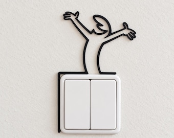 May include: A black line-art decal of a figure with arms raised in celebration, positioned to wrap around the top and left side of a white rectangular double light switch. The figure is stylized with simple curves and appears to emerge from the switch.