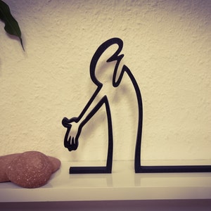May include: Black metal sculpture of a person reaching out with one arm. The sculpture is on a white shelf with two brown stones in front of it.