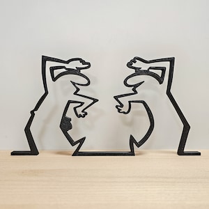 Może przedstawiać: Czarna metalowa rzeźba przedstawiająca dwie postaci stojące naprzeciwko siebie z uniesionymi rękami nad głowami.