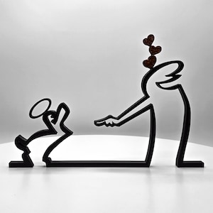 May include: Black metal sculpture depicting two stylized figures. One figure kneels with a halo, facing another figure with outstretched arm and three heart shapes above its head. The sculpture is on a white surface.