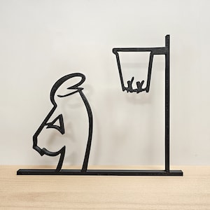 Peut inclure: Silhouette noire d'une personne tirant un ballon de basket dans un panier. La silhouette est en métal et est montée sur une base en bois.