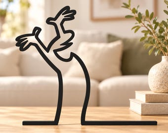 May include: A black metal sculpture featuring a continuous line design that forms the silhouette of a character with raised arms and hands. The object has a flat base and a thin, minimalist profile, standing upright as a decorative tabletop piece.