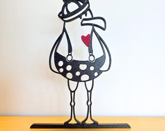 May include: A whimsical black metal sculpture of a bird-like figure wearing a hat and suspenders. The figure has a polka-dot pattern on its body and a red heart on its chest. The sculpture is mounted on a black base.