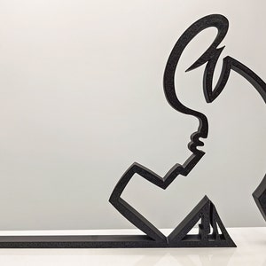 May include: Black abstract sculpture of a person with their head tilted back and their arm raised. The sculpture is made of a single piece of material and is mounted on a black base.