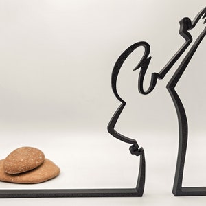 May include: Black metal silhouette of a person with arms raised, standing on a black base. Two brown stones are in front of the sculpture.