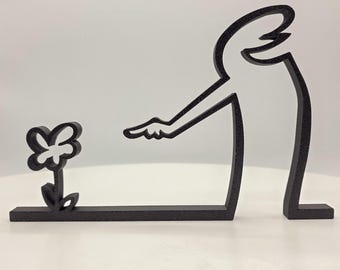 May include: Black metal sculpture of a person pointing to a flower. The sculpture is made of a single piece of metal and is designed to be displayed on a shelf or table.