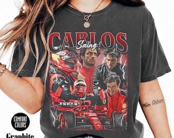 Op de afbeelding: Een antracietgrijs t-shirt met een grafische print van Formule 1-coureur Carlos Sainz. Het ontwerp bevat meerdere afbeeldingen van de coureur in racekleding, een rode racewagen en bliksemschichten, met de tekst "CARLOS SAINZ" in rode letters.