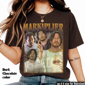 May include: Dark chocolate-colored t-shirt featuring a vintage-style graphic of Markiplier. The design includes multiple images of Markiplier with the name "MARKIPLIER" in gold lettering. The text "up 2-3 size for Oversized" is also visible.