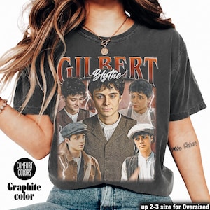 May include: A charcoal gray short-sleeve t-shirt featuring a graphic print of a young man in various poses and outfits. The text GILBERT is printed in large orange letters at the top, with the name Blythe in white script below it.