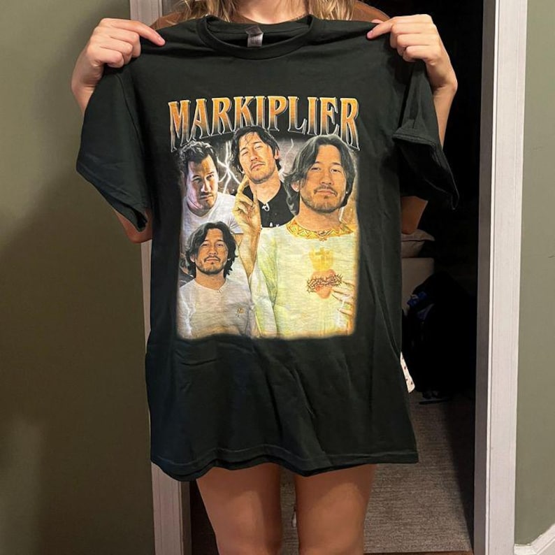 May include: Dark green t-shirt featuring the name "MARKIPLIER" in gold lettering above a collage of four images of a man. The images are in a vintage style, with a faded appearance.