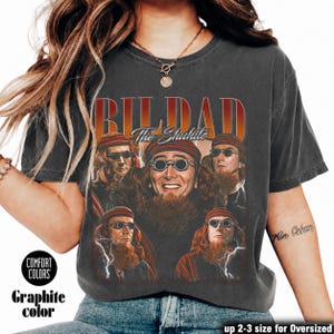 May include: A charcoal gray short-sleeve t-shirt featuring a graphic print of a man with a beard and round glasses in multiple poses. The text BILDAD and The Shuhite is printed in a bold, distressed font above the images, accented by white lightning.