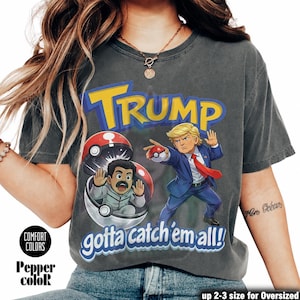 Può includere: T-shirt grigio scuro con una grafica con la scritta "TRUMP" in giallo e blu, e illustrazioni di cartoni animati di due figure in un design a tema Pokémon. È presente anche il testo "gotta catch 'em all!".